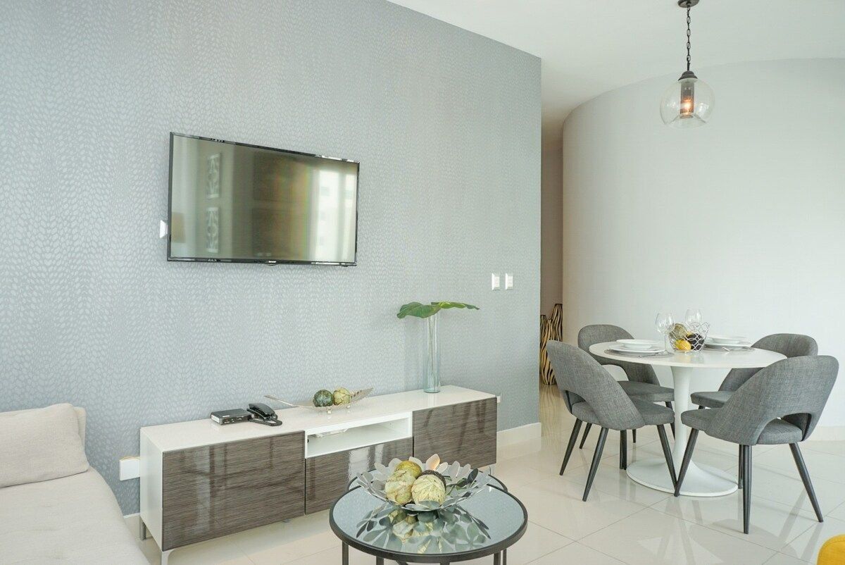 Una sala moderna con un televisor de pantalla plana montado en la pared, una consola blanca con un florero y adornos, una mesa de comedor redonda con sillas grises y una mesa de centro de vidrio con flores.