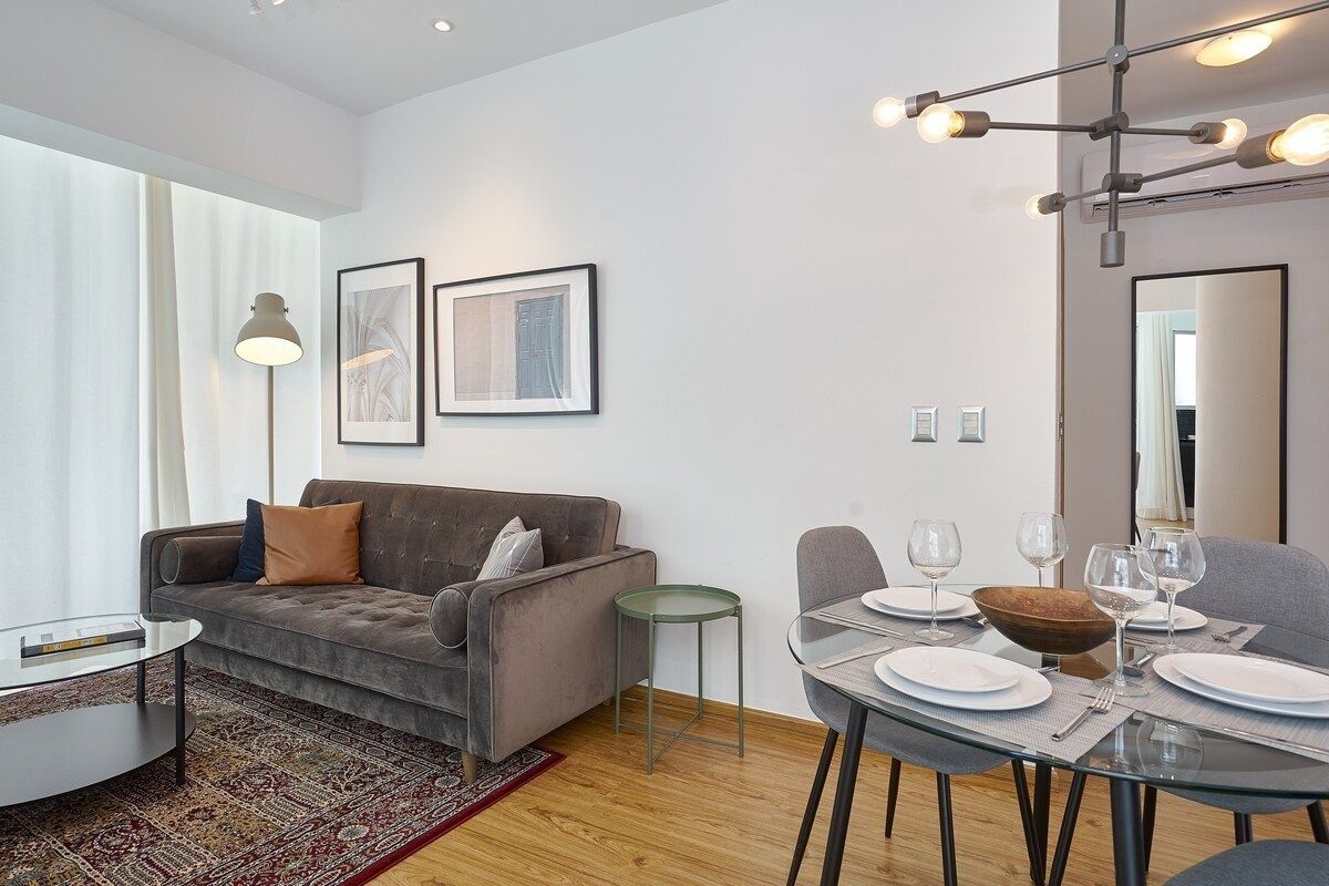 Een moderne woonkamer met een grijze bank, een glazen salontafel en een eethoek met borden en glazen.