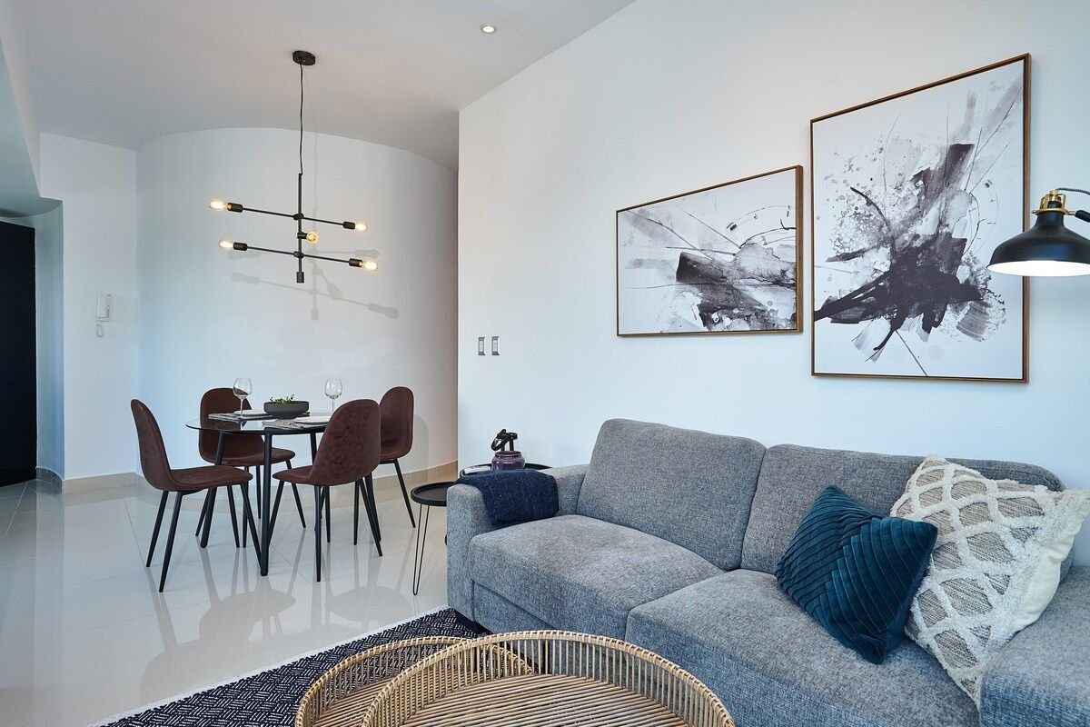 Una sala moderna con un sof á gris, una mesa de comedor con sillas y arte abstracto en la pared.