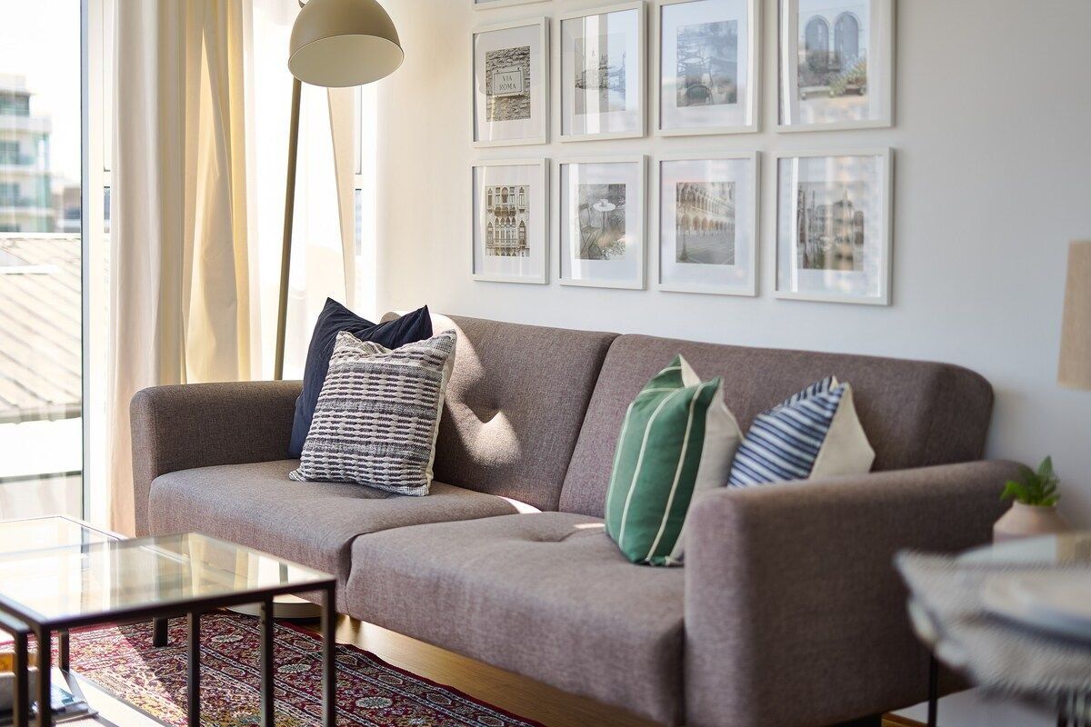 Un salon moderne avec un canapé gris, de nombreux coussins, une table basse en verre et des tableaux encadrés accrochés au mur.