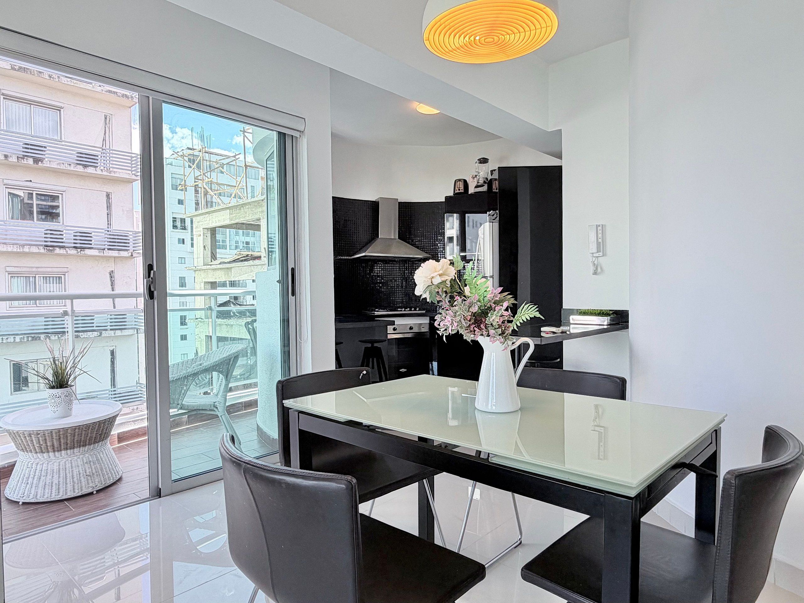 Une cuisine moderne avec un plan de travail noir, un vase blanc orné de fleurs et un coin repas comprenant une table en verre et des chaises noires.