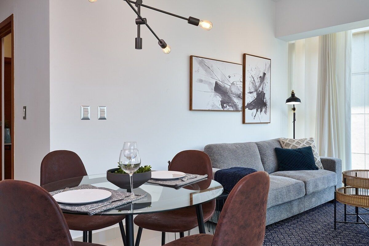 Een moderne woonkamer met een grijze bank, een eettafel met stoelen en twee abstracte schilderijen in lijsten aan de muur.