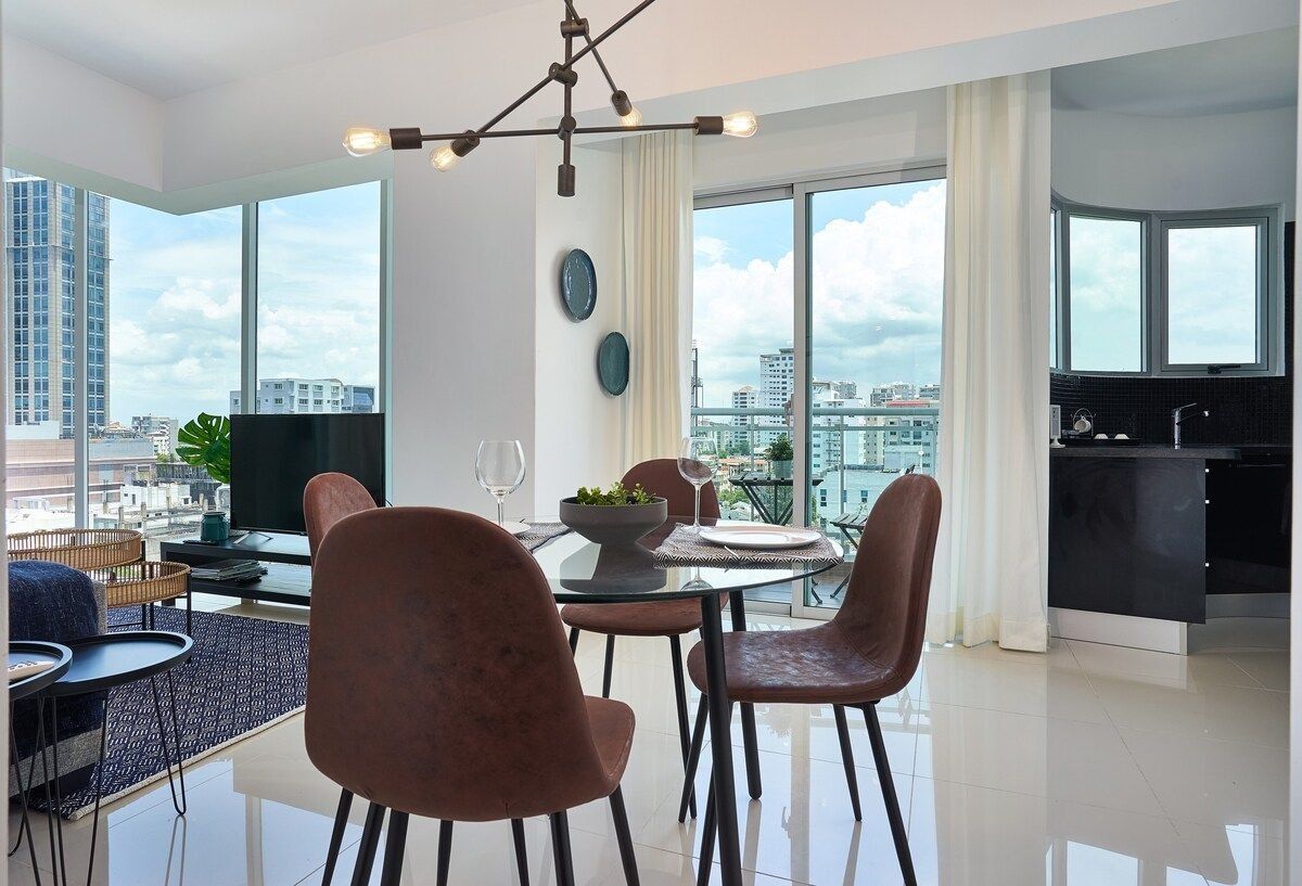A modern dining area with a glass table, four chairs, a bowl of fruit, and a city view.