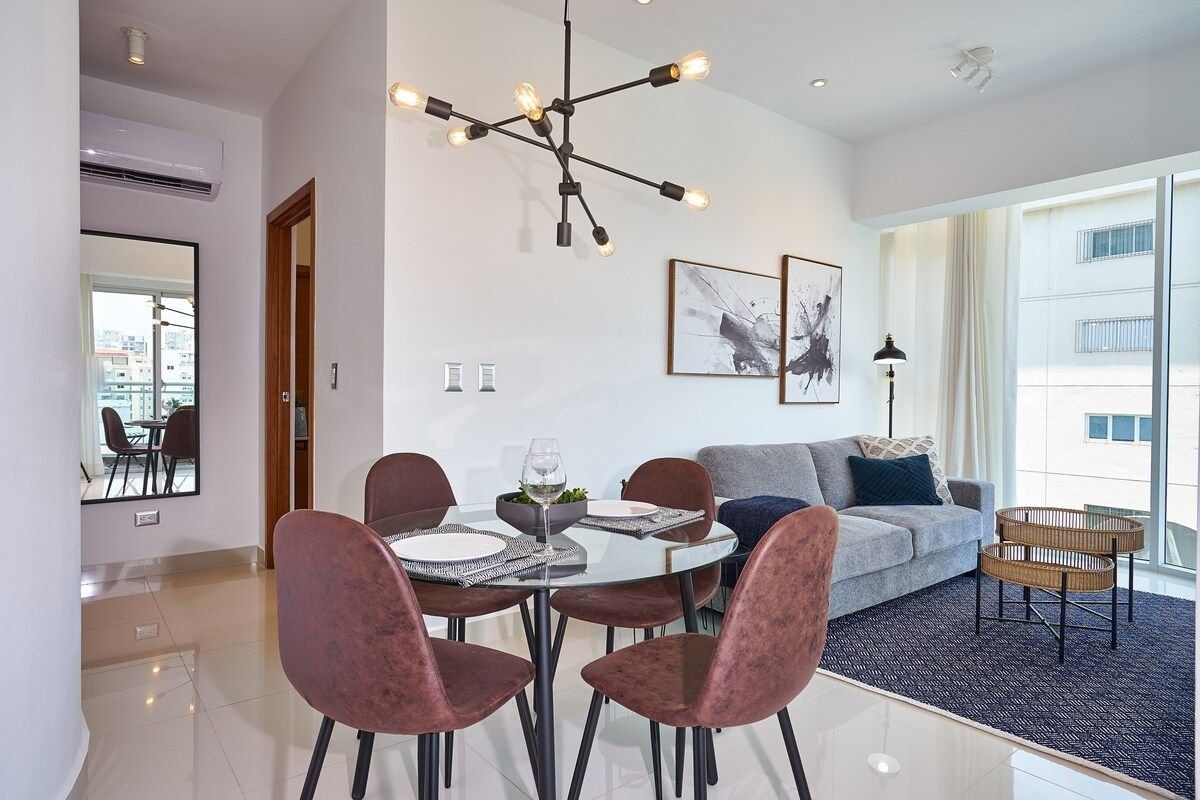 A modern living room with a dining area, featuring a chandelier, a sofa, and a view of the city.