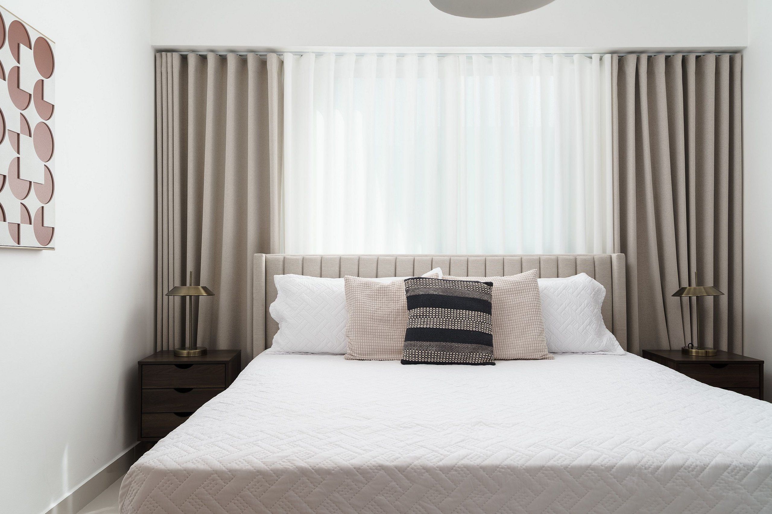 Una cama bien tendida con colcha blanca, dos mesitas de noche con lámparas y un ventanal con cortinas translúcidas.
