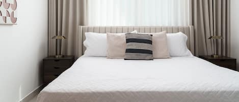 A neatly made bed with a white quilt, two bedside tables with lamps, and a large window with sheer curtains.