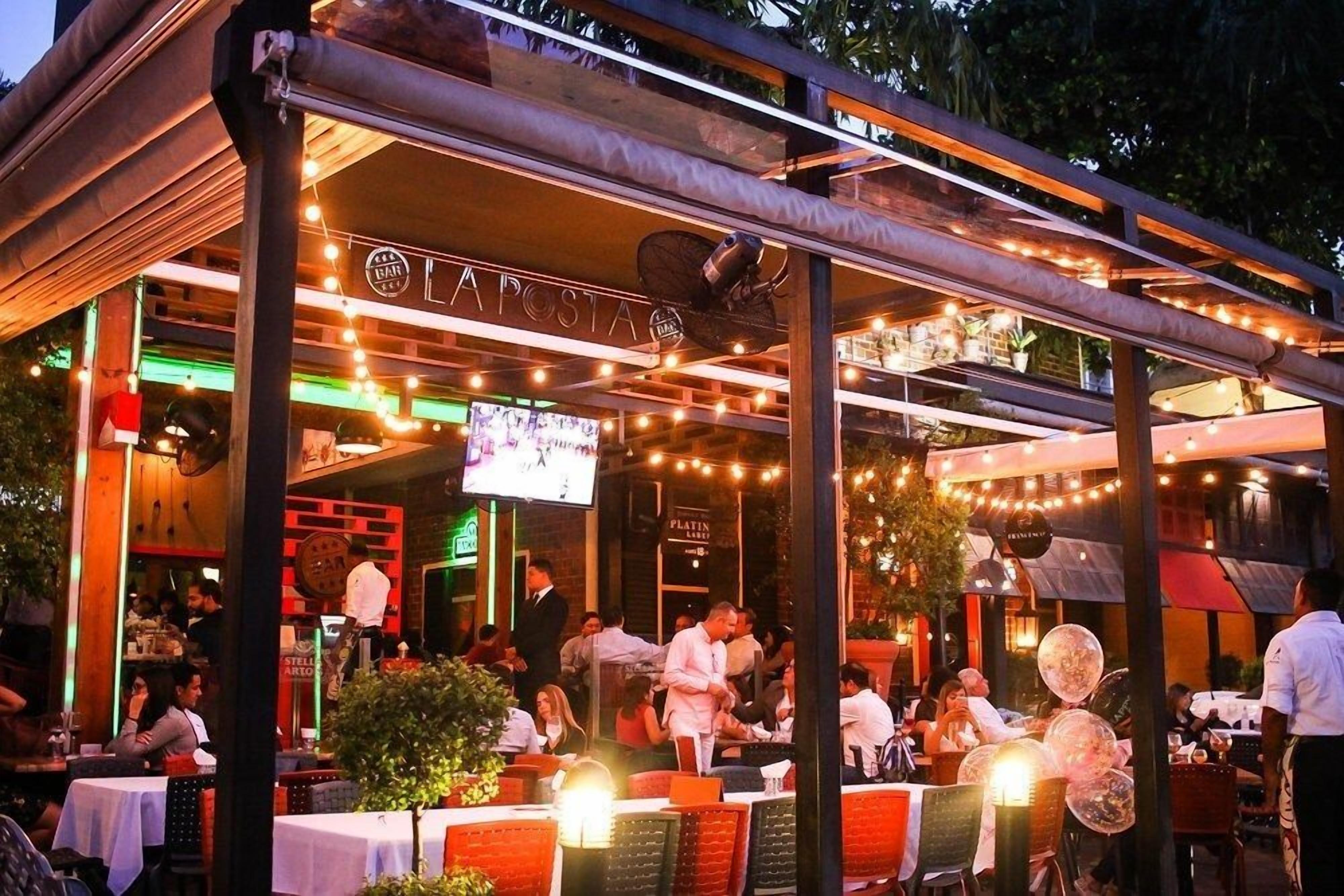 An outdoor dining area with tables and chairs, a bar area, and a sign that reads "APOSTA."