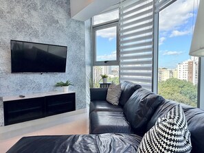 A modern living room with a black leather sofa, a mounted TV, and a city view through large windows. - Torre Armonia 24 (Santo Domingo)