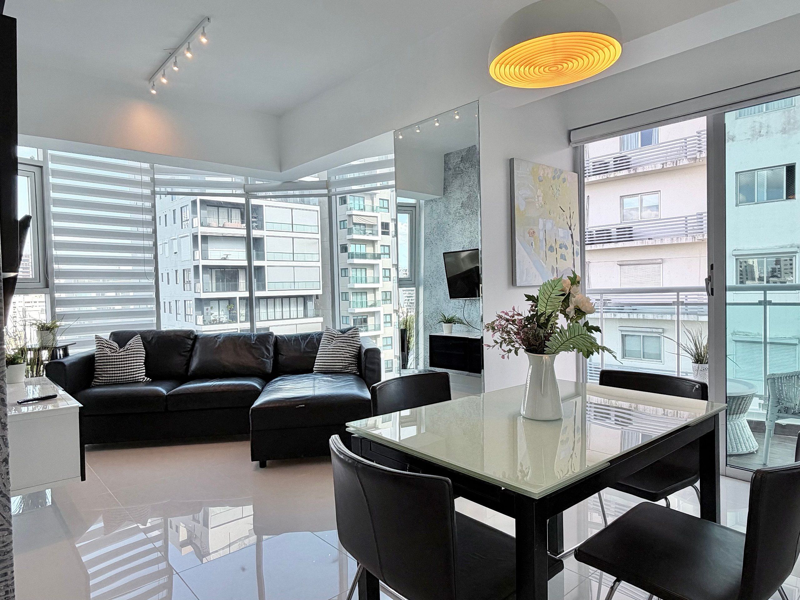 Una sala de estar moderna con un sofá de cuero negro, una mesa de comedor de vidrio y un ventanal con vistas a rascacielos.