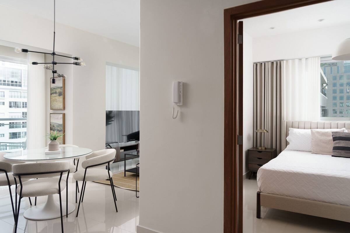A modern hotel room with a bed, bedside table, lamp, and a view of the city.
