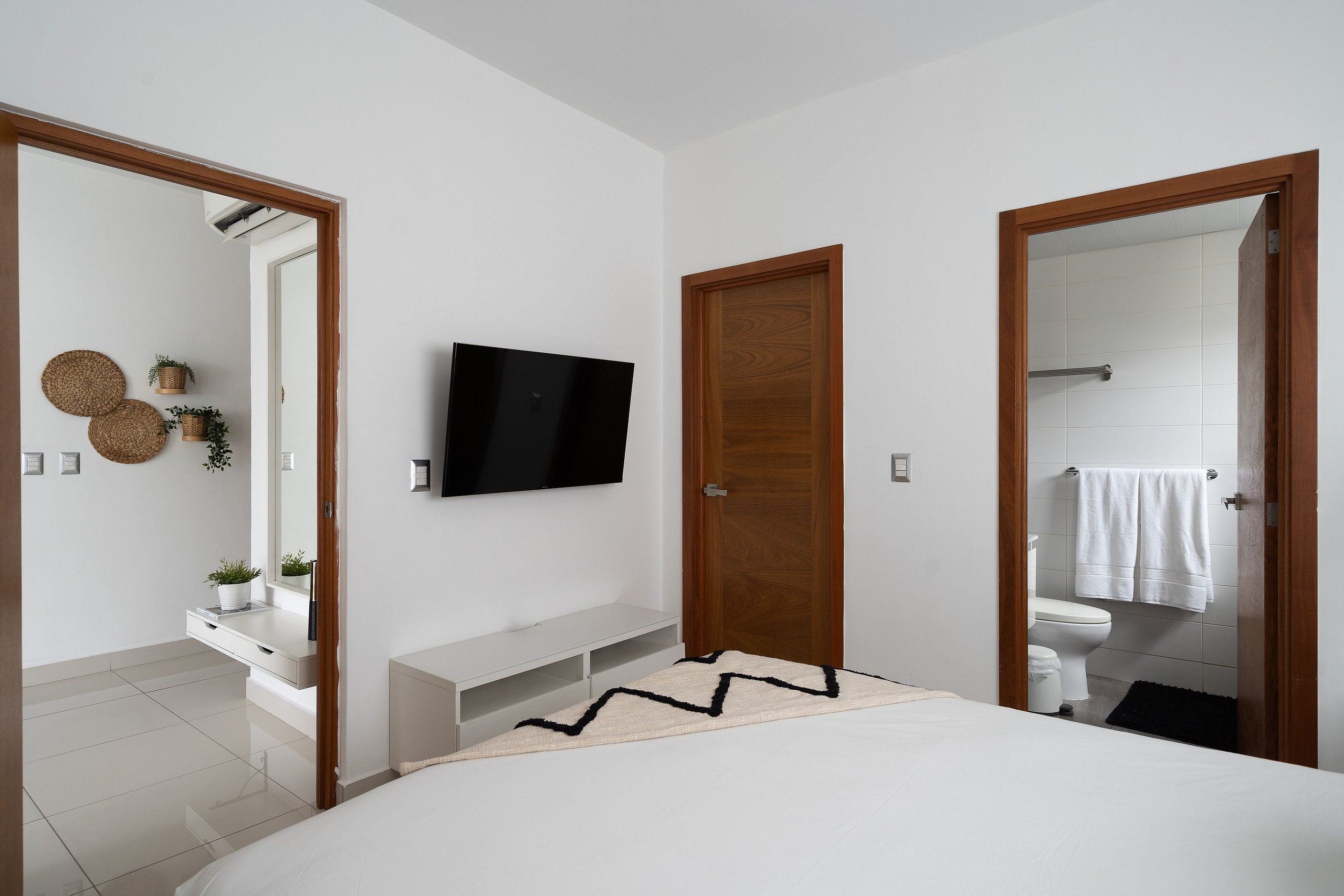 Un dormitorio moderno con cama, televisor montado en la pared, puerta de madera y baño a la vista a través de una puerta abierta.