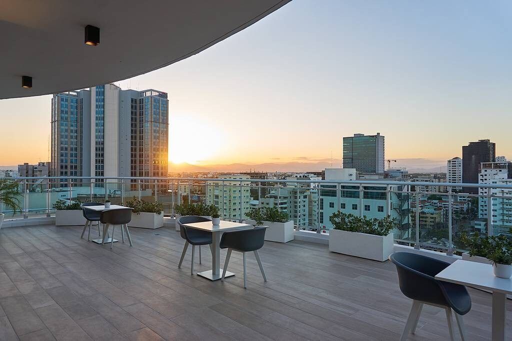 A rooftop terrace with a city view at sunset, featuring a table and chairs, potted plants, and modern buildings in the background.