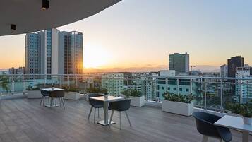 A rooftop terrace with a city view at sunset, featuring a table and chairs, potted plants, and modern buildings in the background.