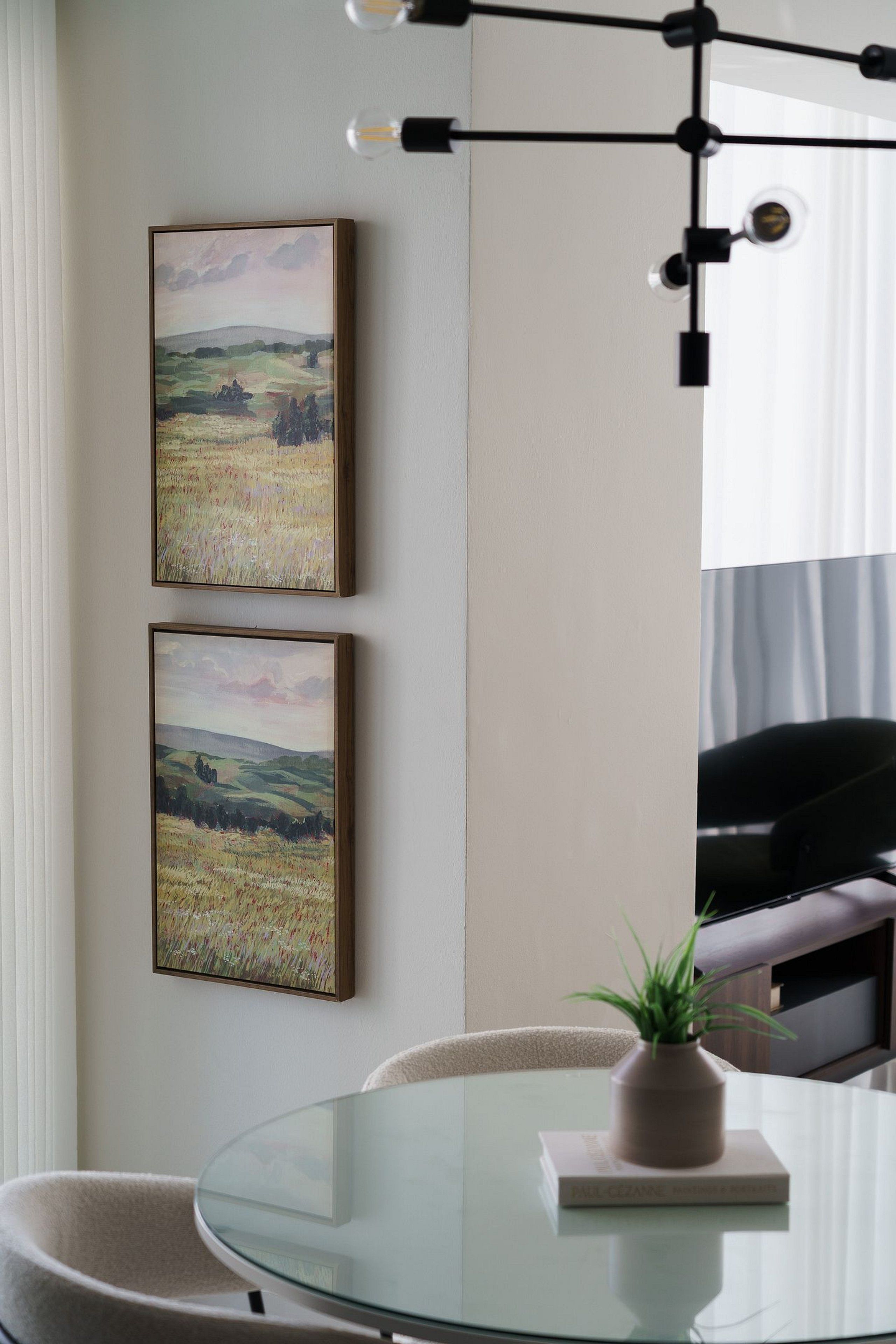 Un espace de salle à manger moderne avec une table en verre, deux tableaux représentant des paysages encadrés, une plante en pot et un luminaire contemporain.