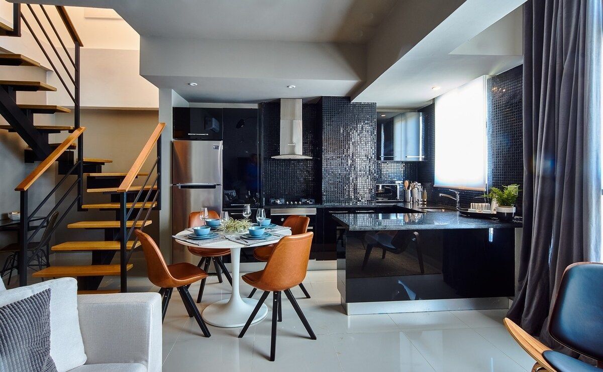 A modern kitchen with a central island, a dining area with a table and chairs, and a staircase with wooden steps and metal railings.