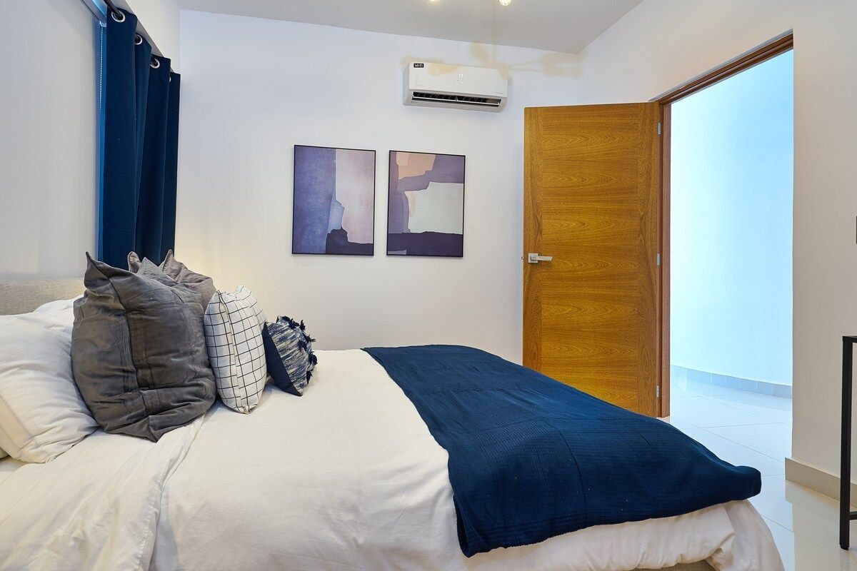 Une chambre à coucher comprenant un lit, des oreillers, une couverture, un appareil de climatisation et deux œuvres d’art murales.