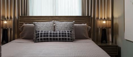 Een net opgemaakt bed met een gewatteerde sprei en twee decoratieve kussens, met aan weerszijden nachtlampjes op houten nachtkastjes.