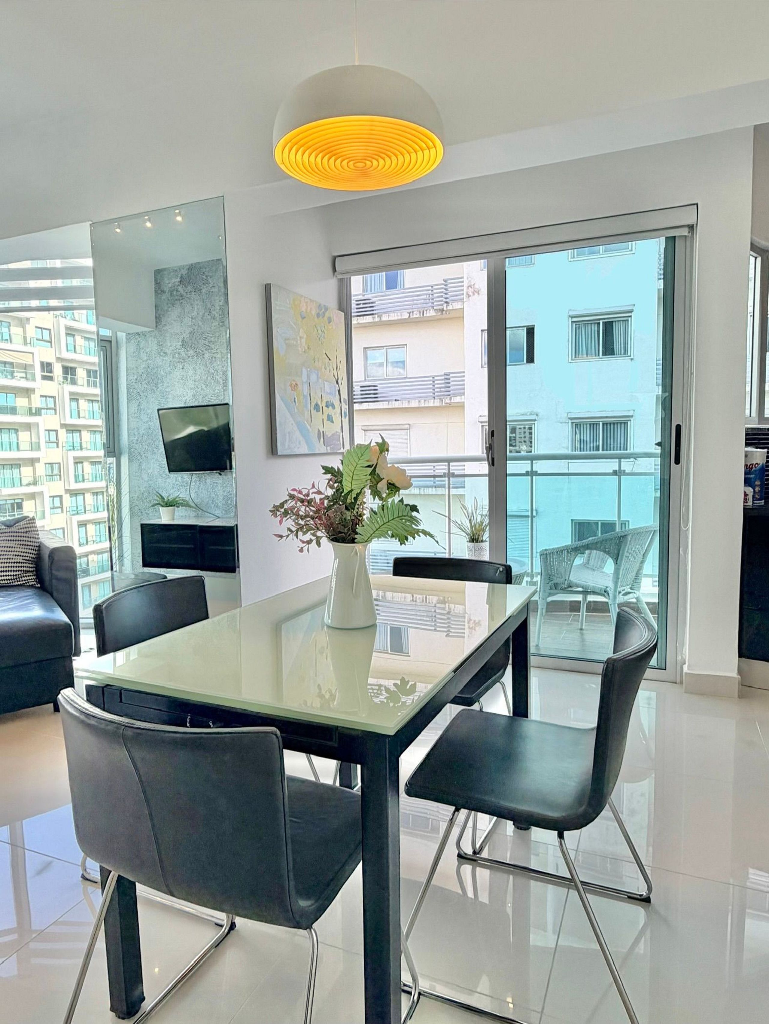 Ein modernes Wohnzimmer mit einem Glastisch, schwarzen Stühlen, einer weißen Vase mit Blumen, einem Wand-Fernseher und einem Balkon mit Blick auf Gebäude.