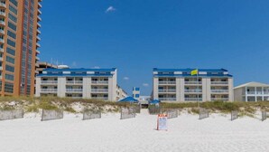 A beachfront hotel with a sandy beach, two-story buildings, and a flag.