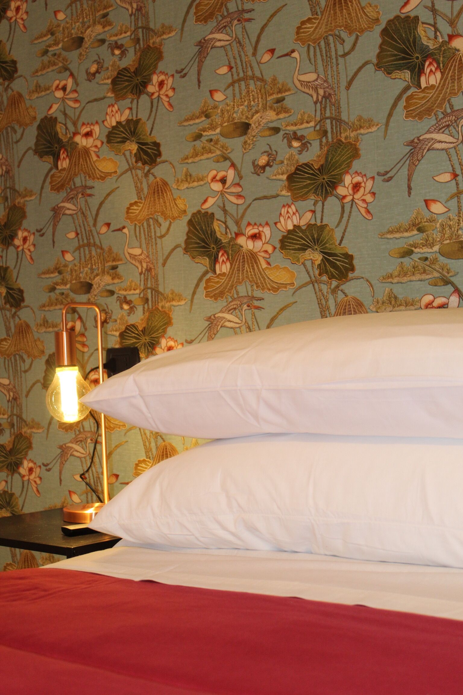 Ein Bett mit weißen Kissen und einer roten Bettdecke, ein Nachttisch mit einer Lampe und ein floraler Wandbehang.