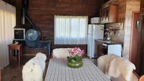 A cozy cabin-style kitchen with wooden walls, a wood-burning stove, a refrigerator, and a dining table with a vase of flowers. - Chale da Montanha (Santo Amaro da Imperatriz)