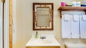A bathroom with a white sink, a wooden cabinet, a mirror with a distressed frame, a toilet, and a shelf with towels and toiletries.