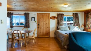 A room with a wooden ceiling, a bed with a blue headboard, a dining table with chairs, and a window with curtains.