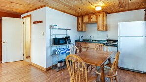 A compact kitchen with wooden cabinets, a microwave, stove, and a refrigerator. There is a dining table with chairs and a small shelf with kitchen items.
