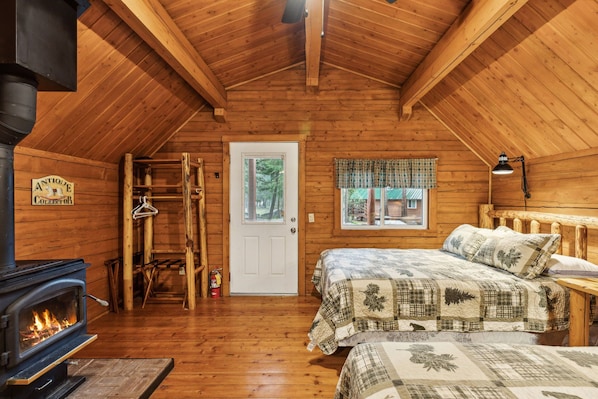 Un dormitorio rústico de cabaña de troncos con una estufa de leña, una cama con colcha, una ventana con cortinas a cuadros y una puerta que da al exterior.