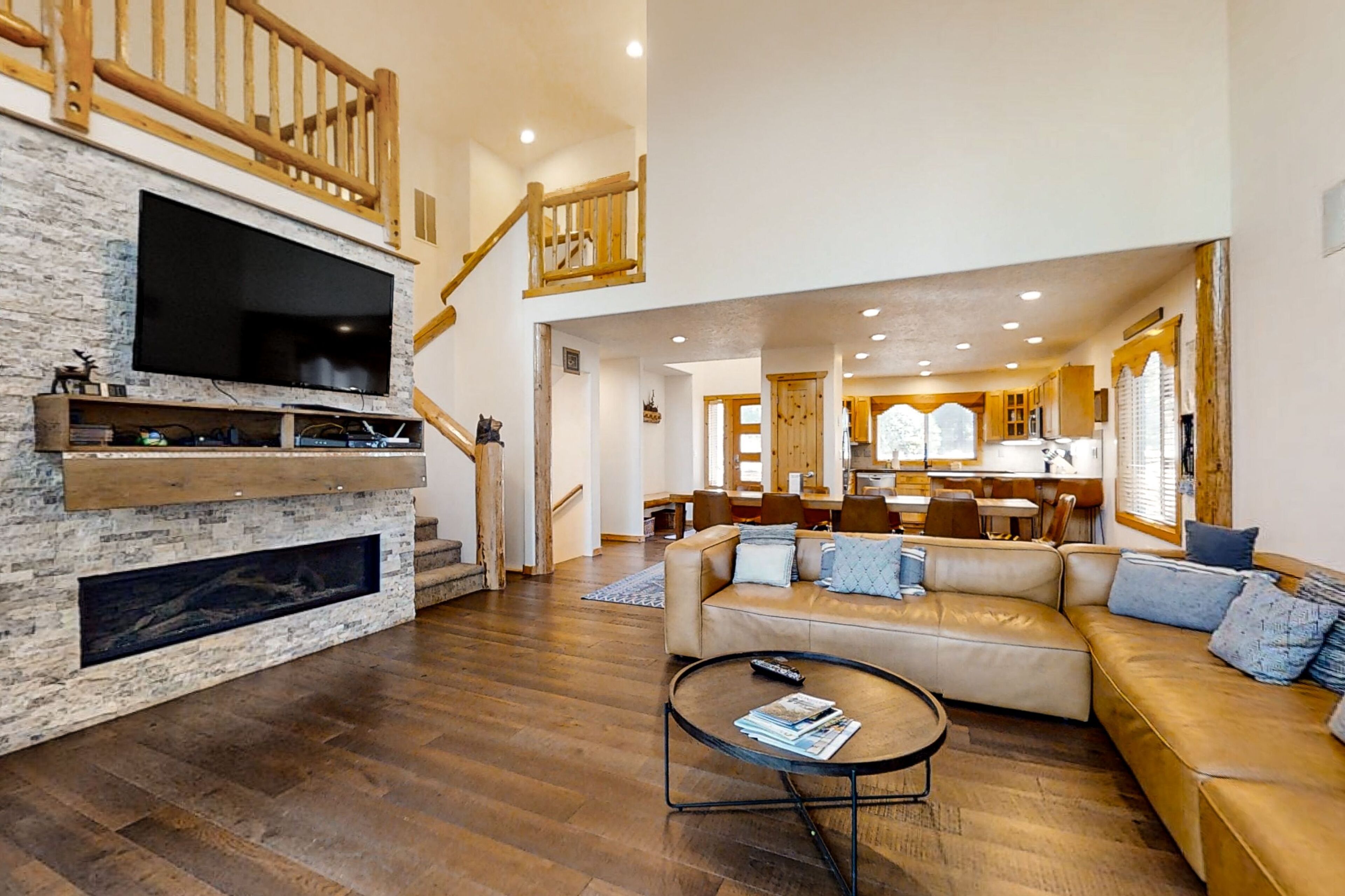 A spacious living room with a stone fireplace, a leather sofa, a coffee table, and a TV mounted on the wall. A staircase leads to an upper level with a wooden railing.