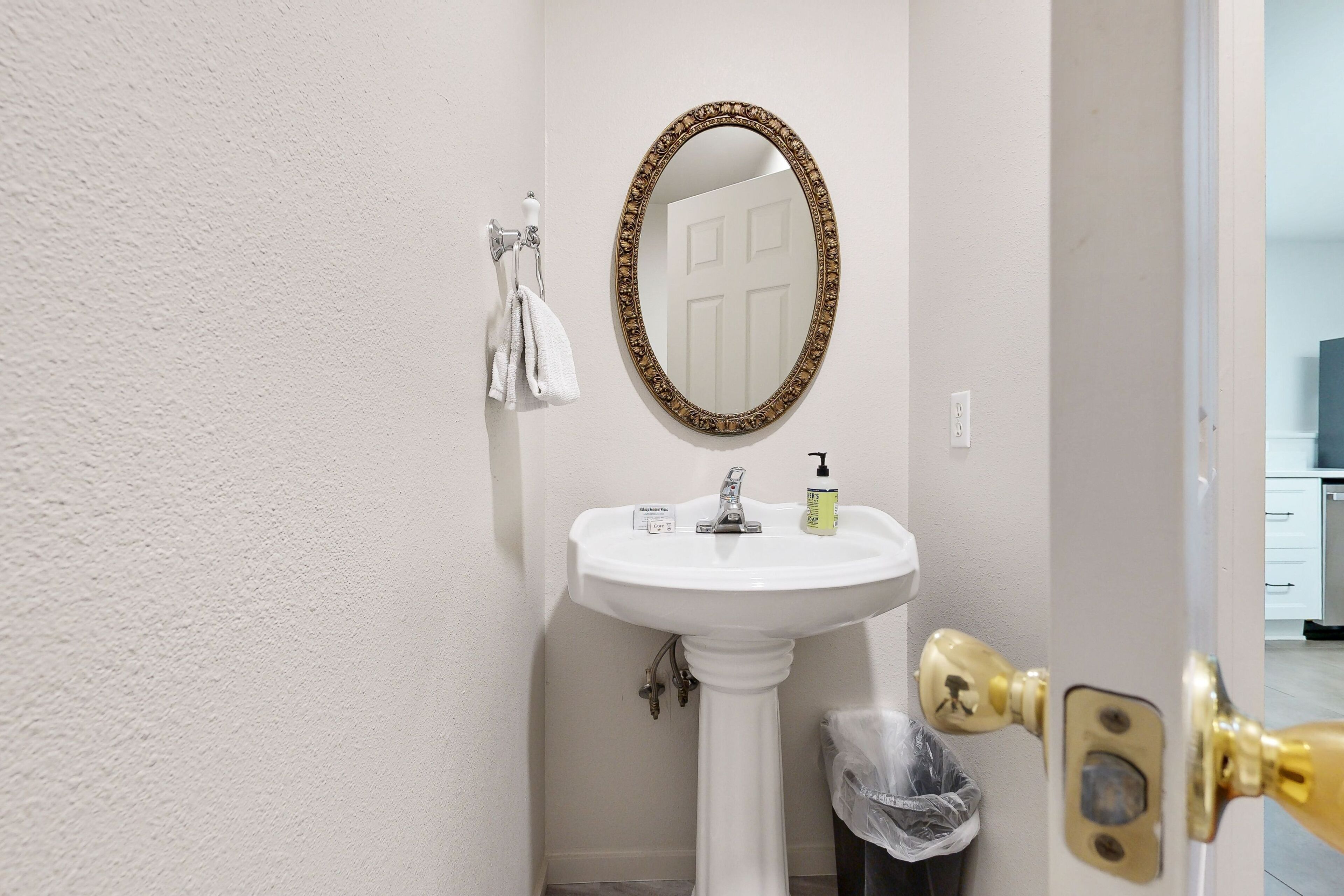 A bathroom with a white pedestal sink, a gold-framed mirror, a towel hanging on a rack, and a door with a brass handle.