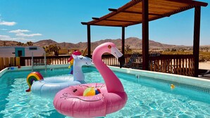 A swimming pool with inflatable swan and unicorn floats, a wooden deck, and a desert landscape in the background. - Stunning 3-Bed/2-Bath Villa with Hot Tub, Pool, & Garden in Joshua Tree (Joshua Tree)