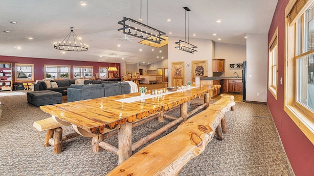 Een ruime woonkamer met een lange houten tafel en banken, een gezellige zithoek met sofa’s, en een keuken op de achtergrond.