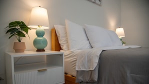 A neatly made bed with white bedding, a bedside table with a lamp, and a potted plant. - Modern Lakeside Escape | Prime Location + Parking (Traverse City)