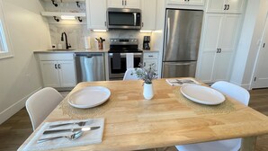 A modern kitchen with white cabinets, stainless steel appliances, and a wooden dining table set for two. - Modern Lakeside Escape | Prime Location + Parking (Traverse City)