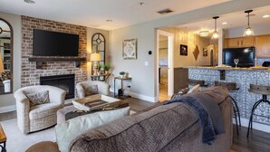 A living room with a brick fireplace, a flat-screen TV, and a kitchen area with a bar. - Cumberland River Views, Walk to Broadway, Downtown & Stadium Rockin' Riverfront (Nashville)