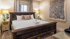 A bedroom with a wooden bed, two bedside tables with lamps, a window with curtains, and a large floral painting on the wall. - Cumberland River Views, Walk to Broadway, Downtown & Stadium Rockin' Riverfront (Nashville)