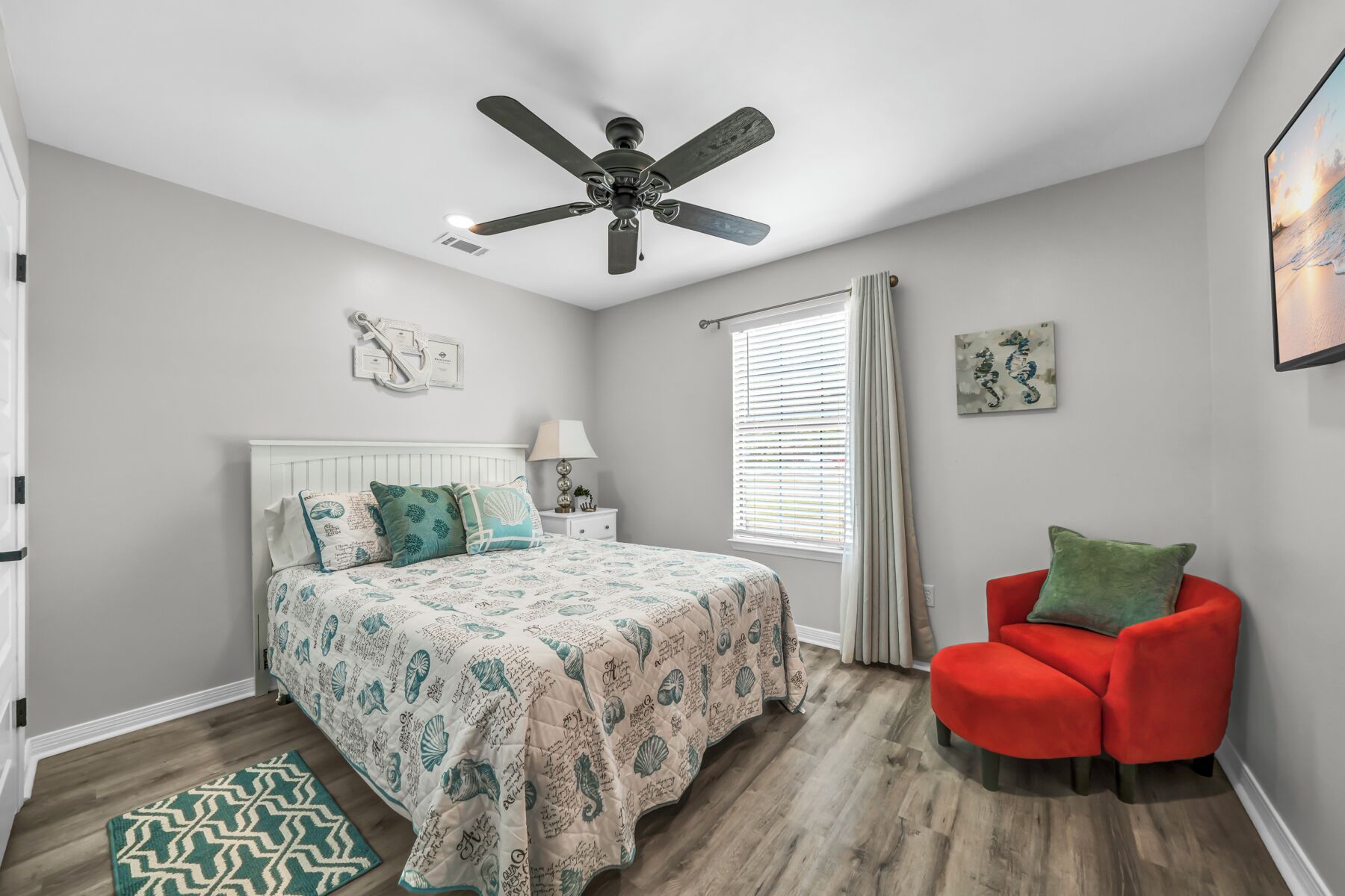 A bedroom with a bed, a red chair, a ceiling fan, a window with blinds, and artwork on the walls.