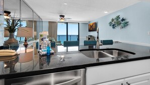 A modern kitchen with a black countertop, stainless steel sink, and white cabinets. There is a ceiling fan, a large window with a view, and a dining area with teal chairs. - Sundestin 1403 (Destin)