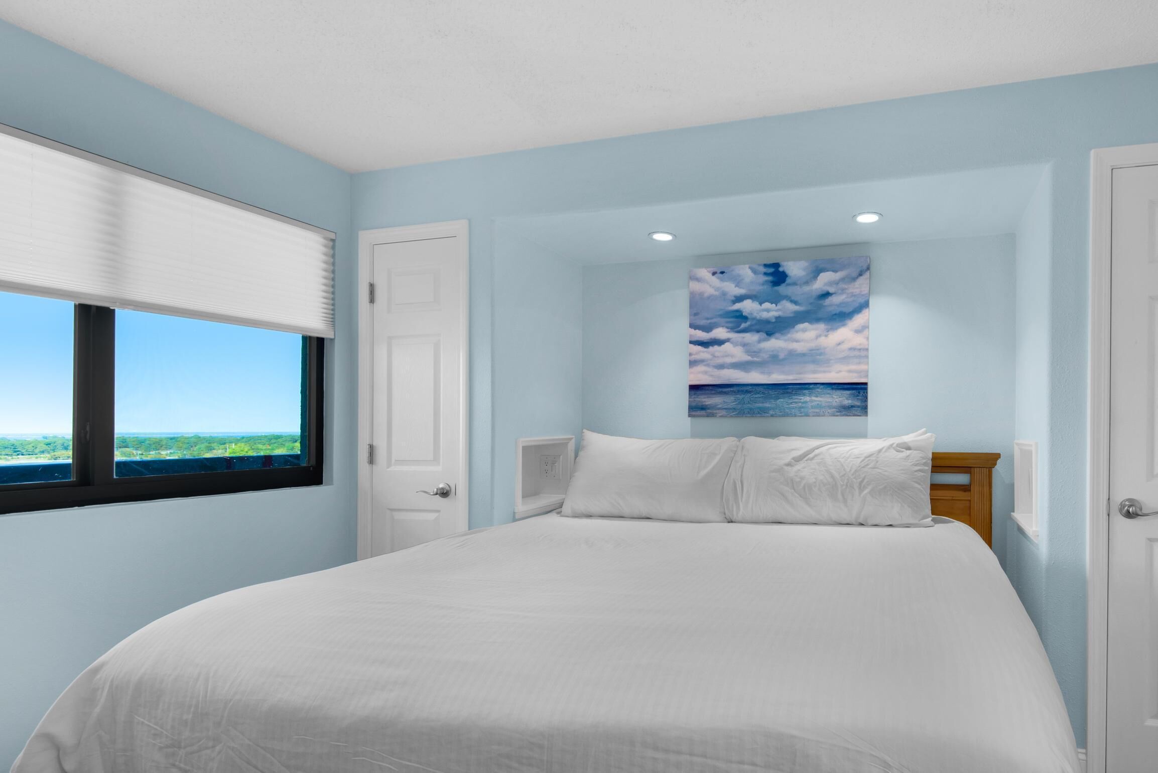 A bedroom with a large bed, a window with blinds, a door, and a painting of a sky and clouds on the wall.