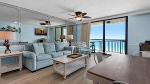 A modern living room with a light blue sofa, a wooden coffee table, and a dining table. There are ceiling fans, a large window with a view of the ocean, and a balcony with a railing. - Sundestin 1403 (Destin)