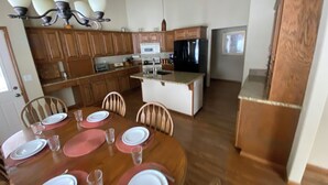 On aperçoit un coin repas avec une table en bois dressée pour quatre personnes, entourée de chaises en bois. Au fond, on distingue une cuisine avec des placards en bois et un réfrigérateur.