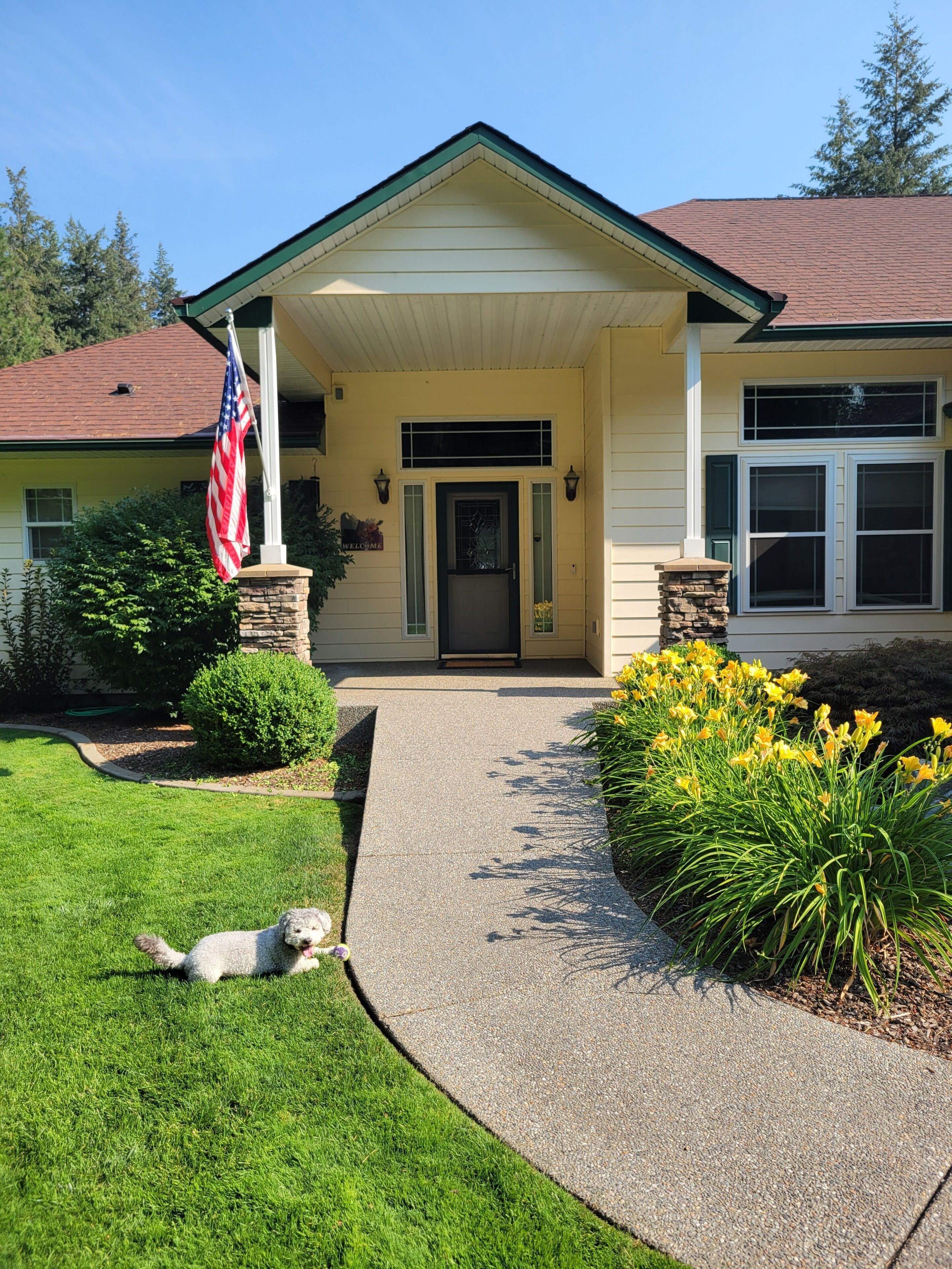 A residential house with a front porch, a flag, and a dog lying on the grass.