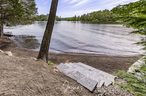 Eine ruhige Uferlandschaft mit Sandstrand, einer Steinsitzbank und einem Baum.
