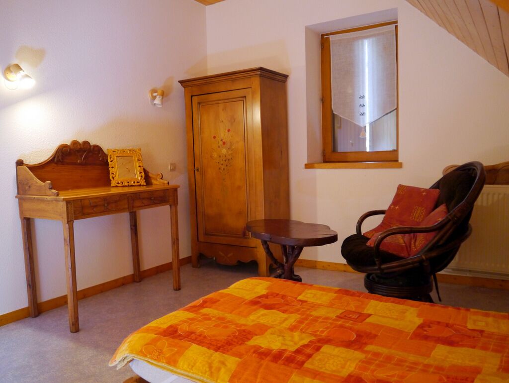 A bedroom with a bed, a wooden wardrobe, a small table, a chair, and a window with a curtain.