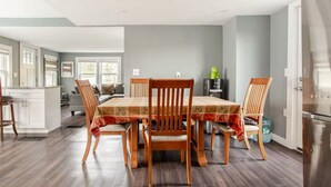 On aperçoit un coin repas avec une table dressée pour quatre personnes, entourée de chaises en bois. Au fond, on distingue une cuisine équipée d’un réfrigérateur et d’un distributeur d’eau.