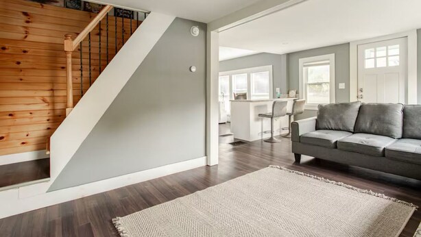 Un salon moderne avec un escalier en bois, un canapé gris et un coin cuisine à l’arrière-plan.