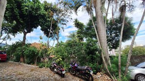 A cobblestone street lined with parked motorcycles and a red car, surrounded by lush greenery and tall trees.