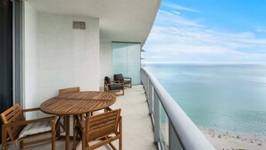 Un balcon avec une table et des chaises en bois, une porte vitrée et une vue sur l’océan.