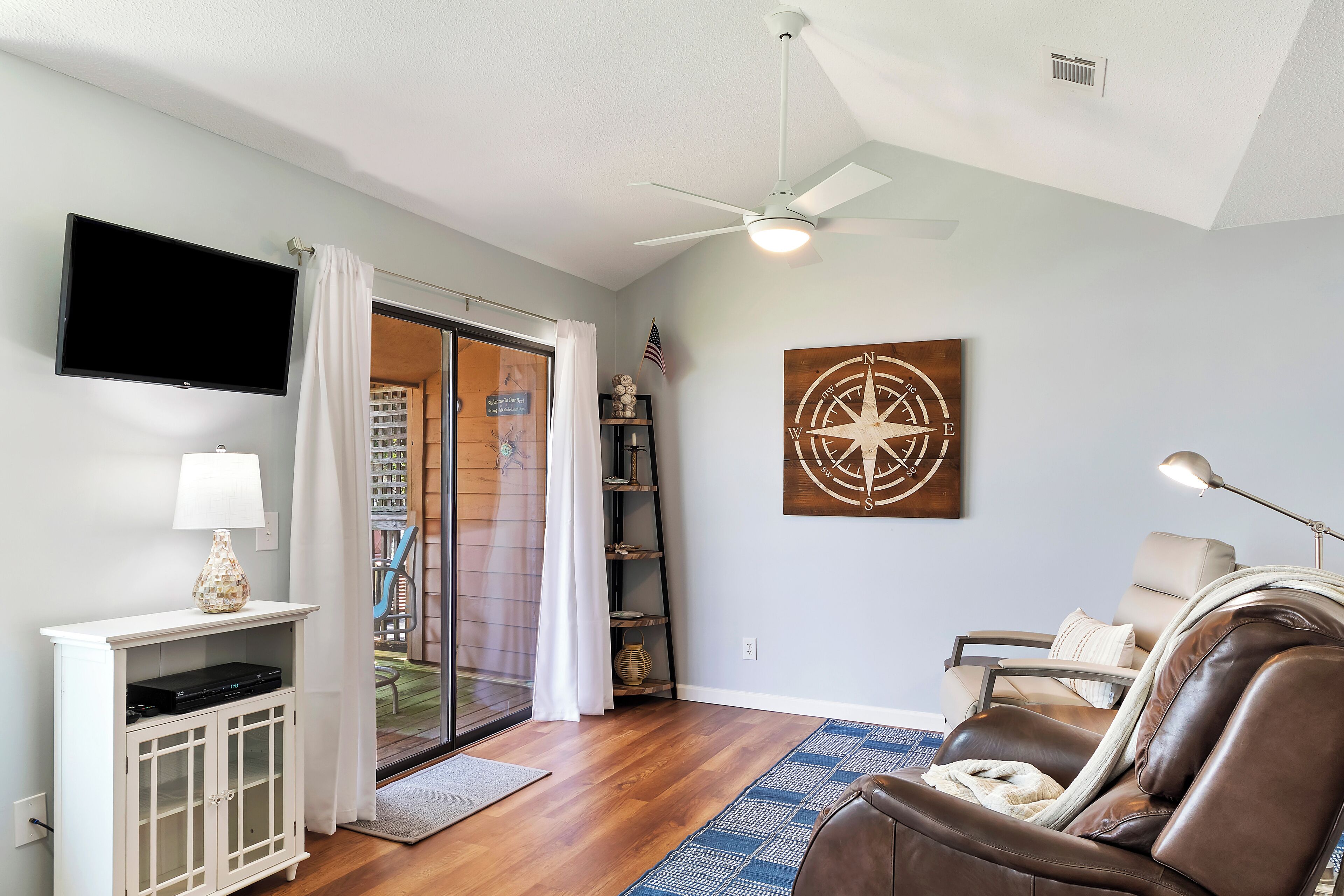 A living room with a flat-screen TV, a ceiling fan, a leather sofa, and a large window with a view of a deck.